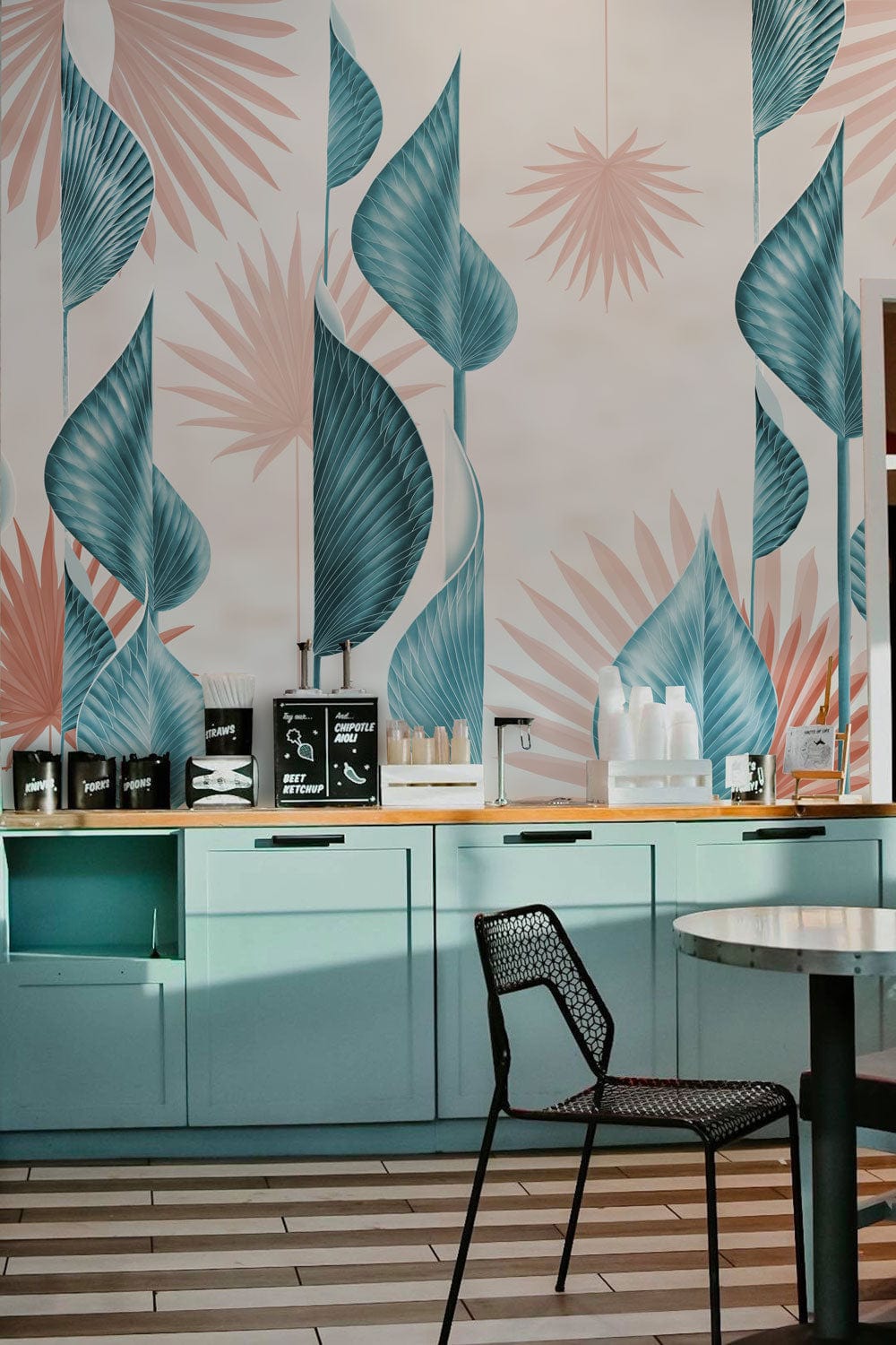 Tropical blue and coral leaf wallpaper in modern kitchen with light blue cabinets, black chair, tiled flooring.