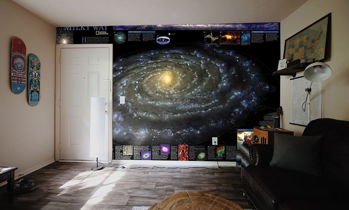 Celestial Galaxy Mural Wallpaper with cosmic blue, golden swirls, dark sofa, and wooden flooring.