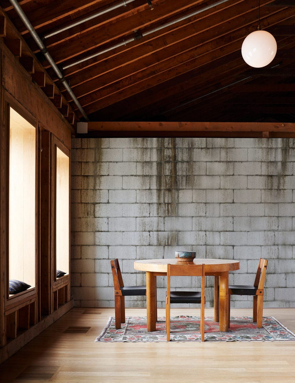 Industrial grey concrete brick wallpaper in loft; wooden table, chairs, wooden ceiling, colourful rug.