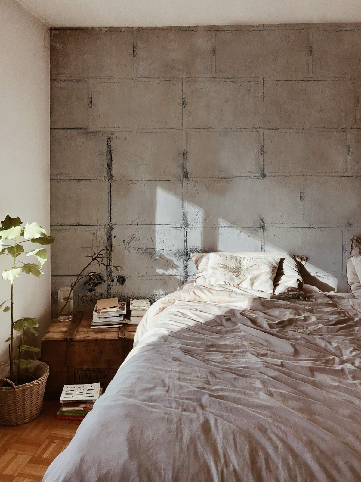Urban bedroom with Industrial Grey Concrete Block Wallpaper, beige bedding, wooden flooring, and natural light.