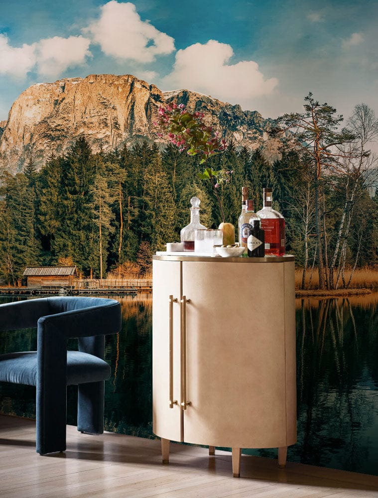 Serene Mountain Lake Reflection Mural Wallpaper with neutral cabinet, dark velvet chair, wooden flooring.