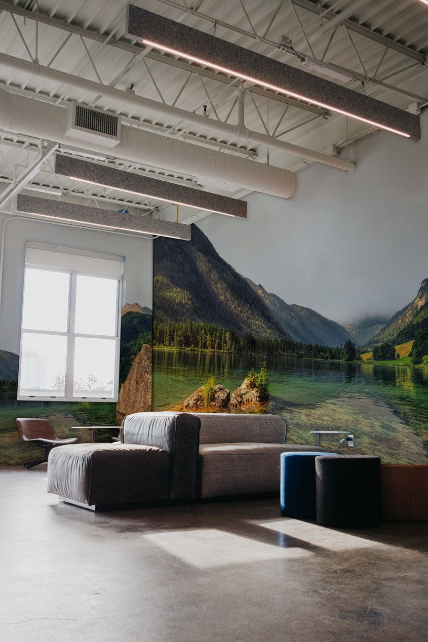 Tranquil mountain lake mural in industrial room, grey sofa, blue stools, concrete flooring visible.