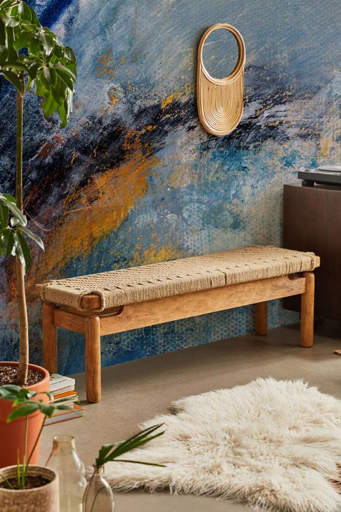 Abstract mural wallpaper with woven bench, round mirror, fluffy rug, and concrete flooring in living area.