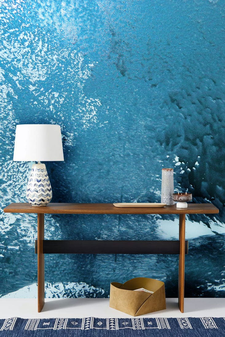 Teal abstract ocean wave mural wallpaper, modern console table, patterned blue rug, ceramic lamp.