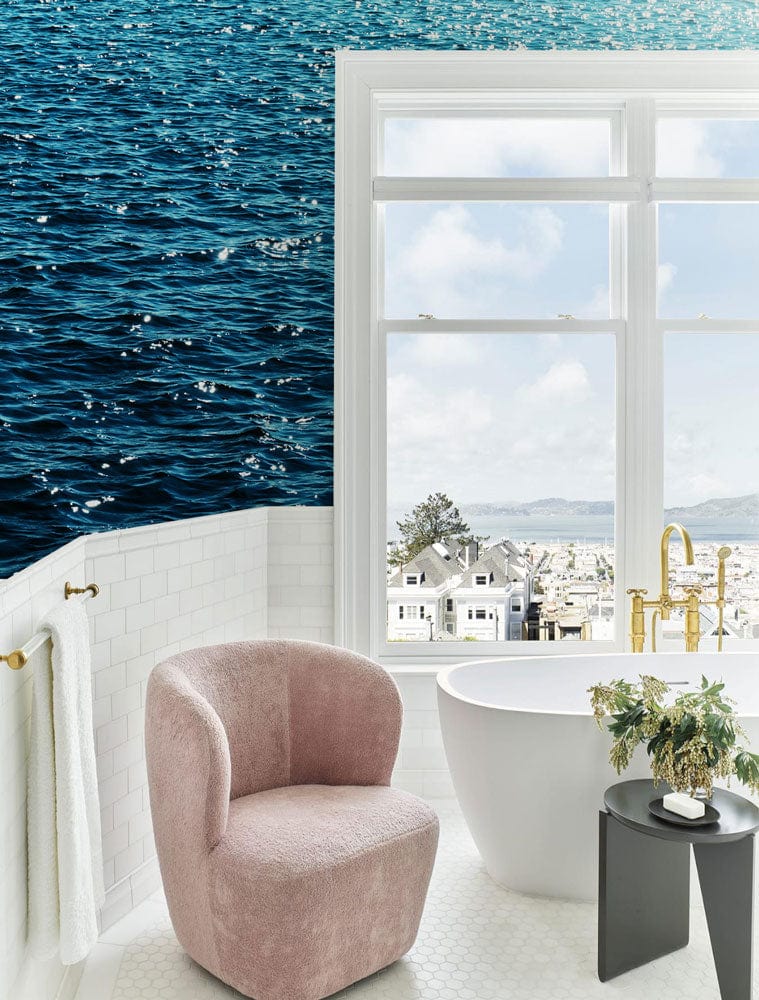 Modern bathroom featuring Tranquil Ocean Blue Ripple wallpaper, white bathtub, pink chair, and elegant decor.