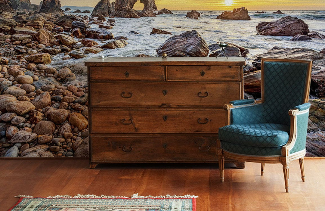 Sunset coastal mural wallpaper, dramatic rocks, wooden dresser, blue vintage chair, hardwood flooring, living room.