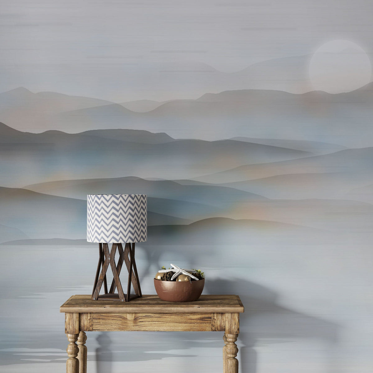 Tranquil misty mountain mural wallpaper in soft blue-grey, rustic table, zigzag lamp, neutral decor.