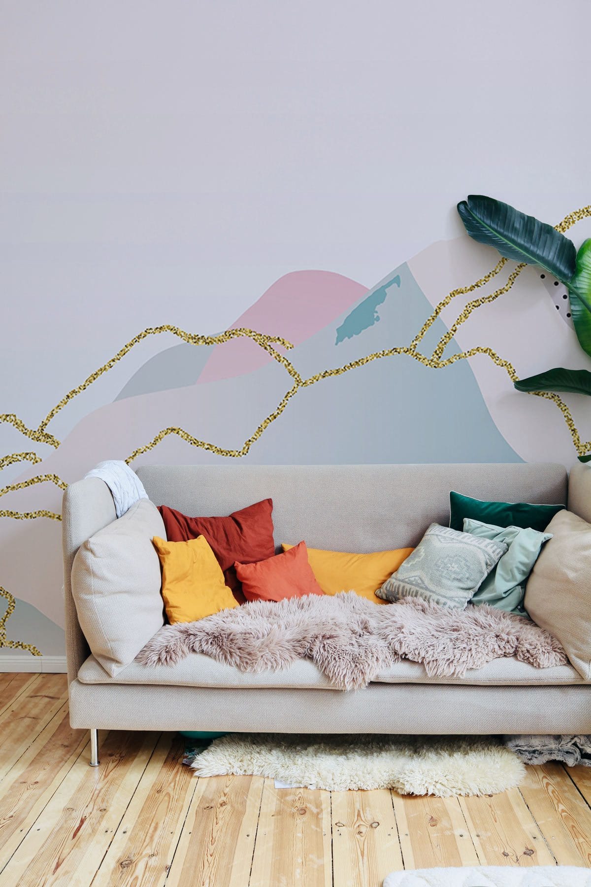 Beige sofa, pastel abstract mountain mural wallpaper, gold accents, wooden flooring, vibrant cushions, living room.