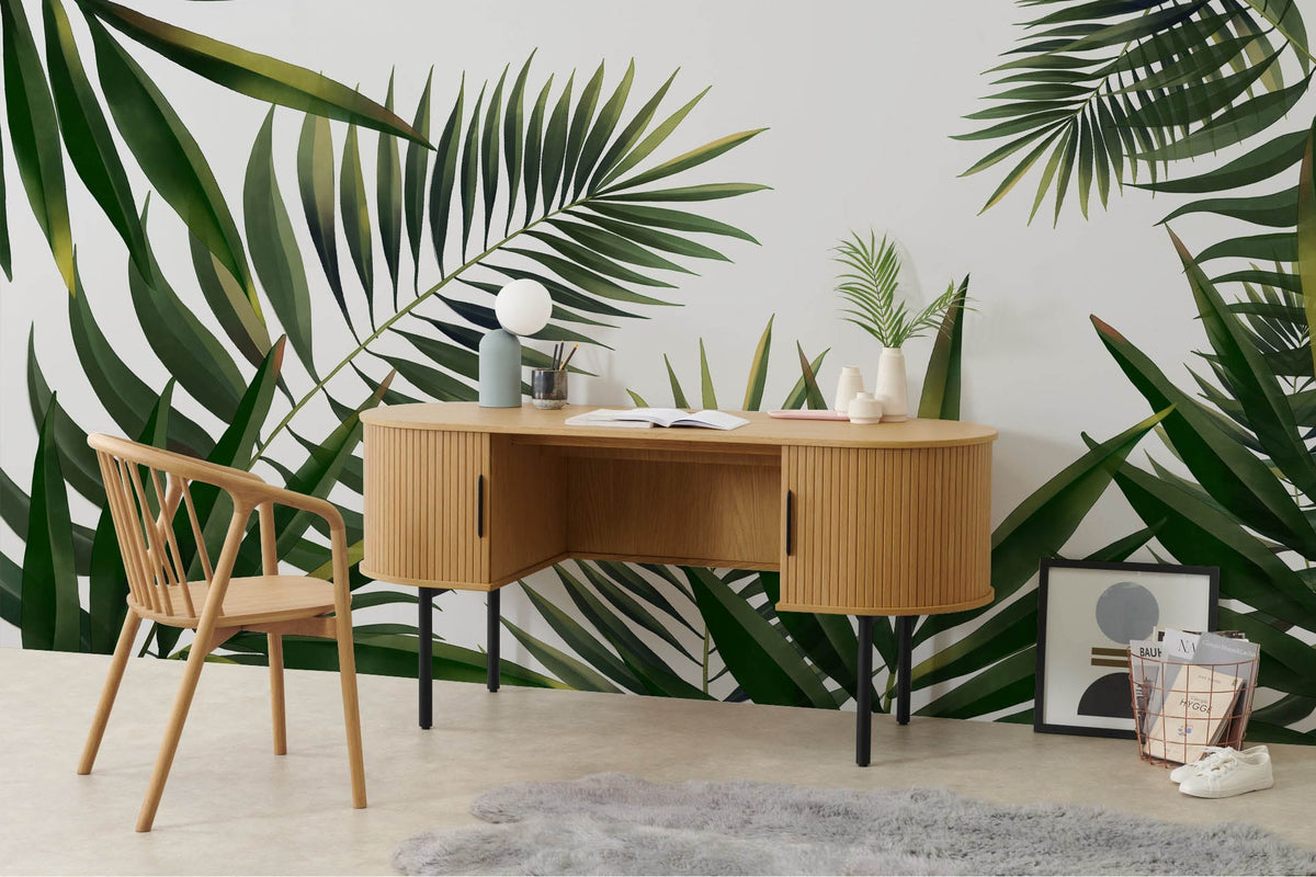 Elegant tropical mural wallpaper with green palm leaves, wooden desk, chair, and grey rug in modern interior.