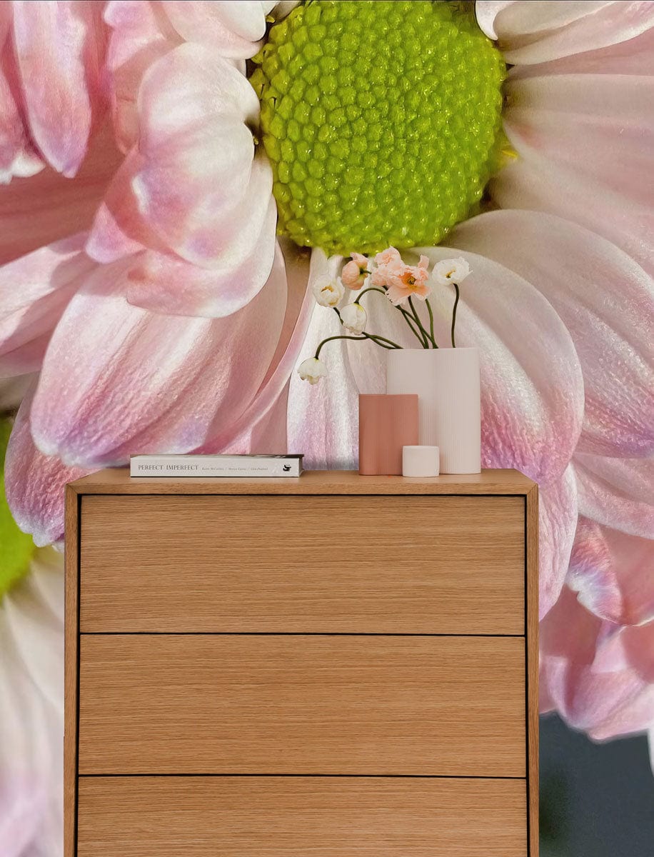 Blush pink daisy mural wallpaper with vibrant green centres, wooden dresser, modern room setting.