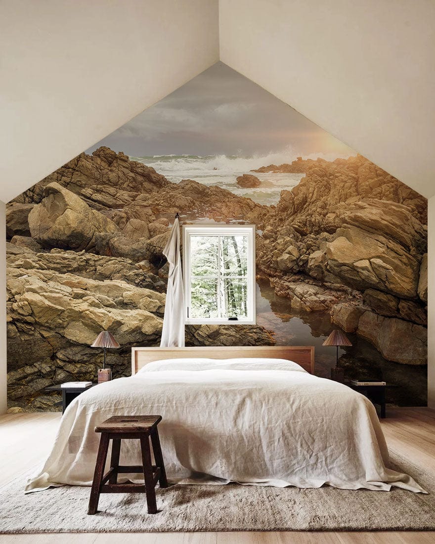 Sunlit Rocky Coastline Mural Wallpaper, bedroom with wooden flooring, minimalist bed and stool.