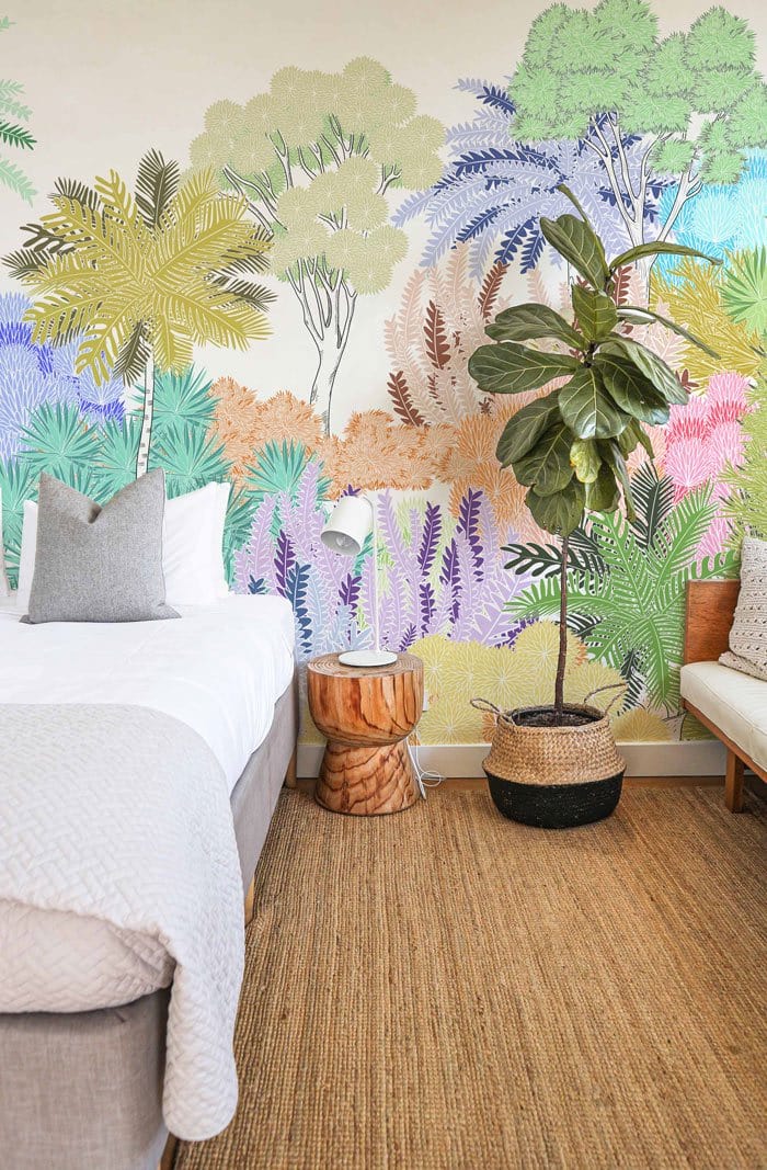 Bedside setting with colourful pastel tropical jungle mural wallpaper, neutral sofa, wooden stool, textured rug.