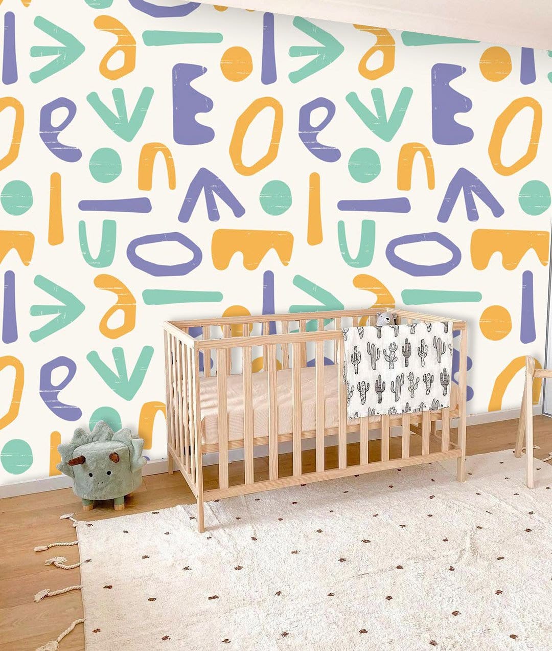 Playful Pastel Geometric Wall Mural in nursery with light wood crib, abstract patterned wallpaper, hardwood flooring.