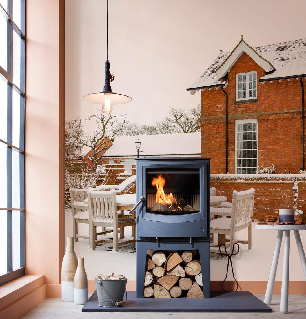 Snowy English Countryside mural wallpaper with brick house, cosy fireplace, and wooden floor in living room.