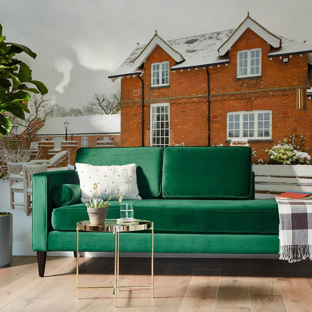 Green velvet sofa, gold side table, woodland scenery, brick house mural wallpaper, wooden floor, plant.