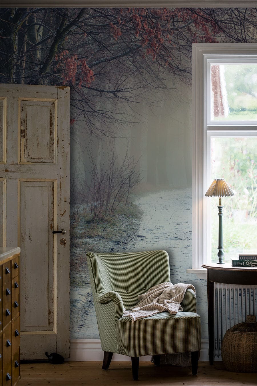 Winter Forest Path Mural Wallpaper featuring serene misty landscape, green armchair, rustic door, table lamp.