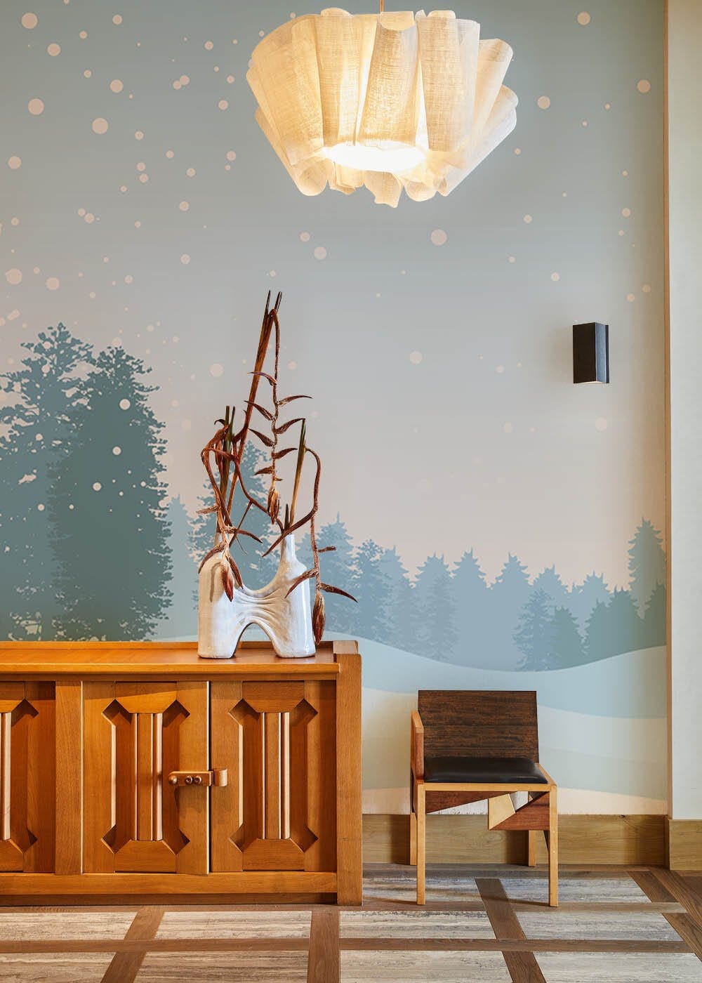 Serene Winter Wonderland Blue Mural Wallpaper with wood sideboard, modern chair, and ambient lighting fixture.