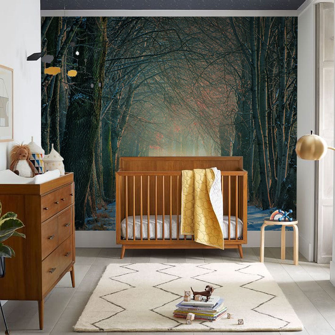 Enchanted Winter Forest Mural Wallpaper in nursery with wooden cot, drawers, beige rug, modern decor.