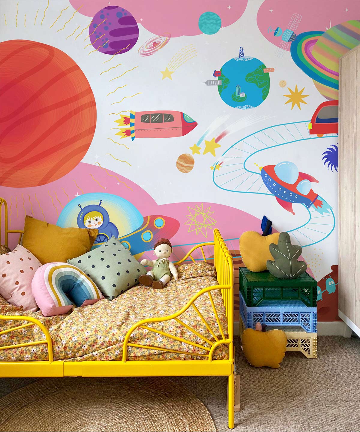 Vibrant Kids' mural in pink space theme, featuring yellow metal bed, cosy cushions, and wooden wardrobe.