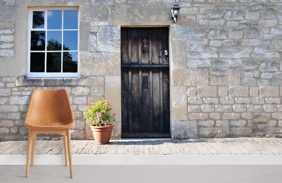 Rustic stone cottage wall mural with wooden door, window, tan leather chair, and grey flooring.