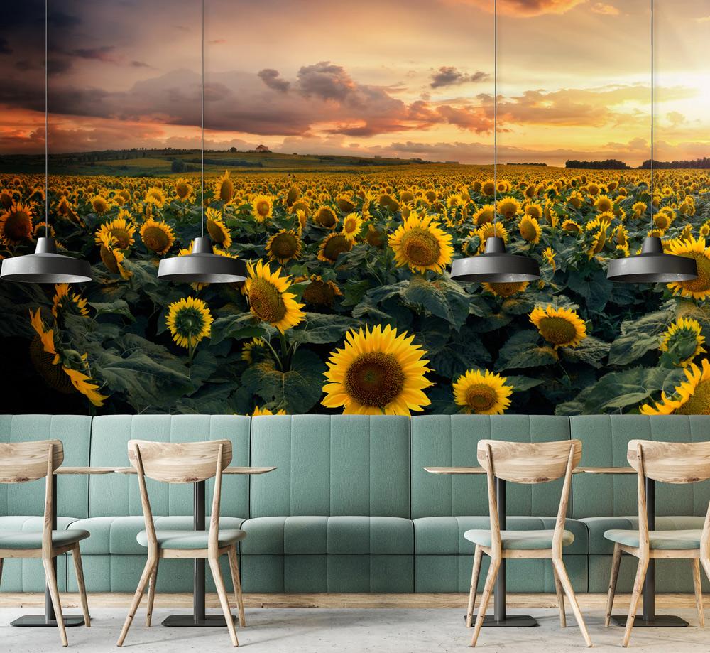 Sunflower sunset mural in modern café with green sofas, wooden chairs, hanging lights, and concrete floor.