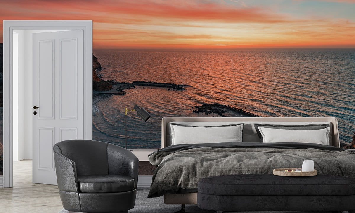 Sunset coastal mural wallpaper in warm tones, grey bed and armchair, neutral flooring, serene bedroom.