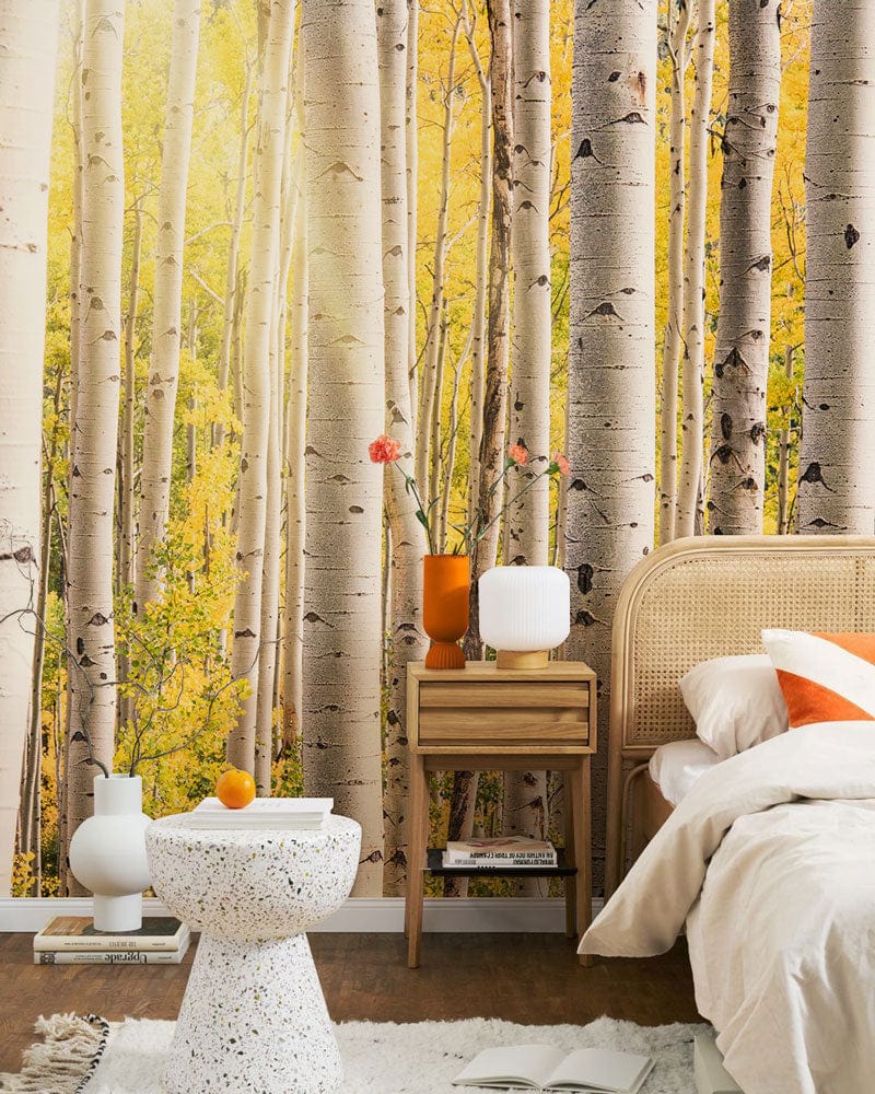 Golden Autumn Birch Forest mural wallpaper in bedroom with beige rattan bed, white terrazzo table.