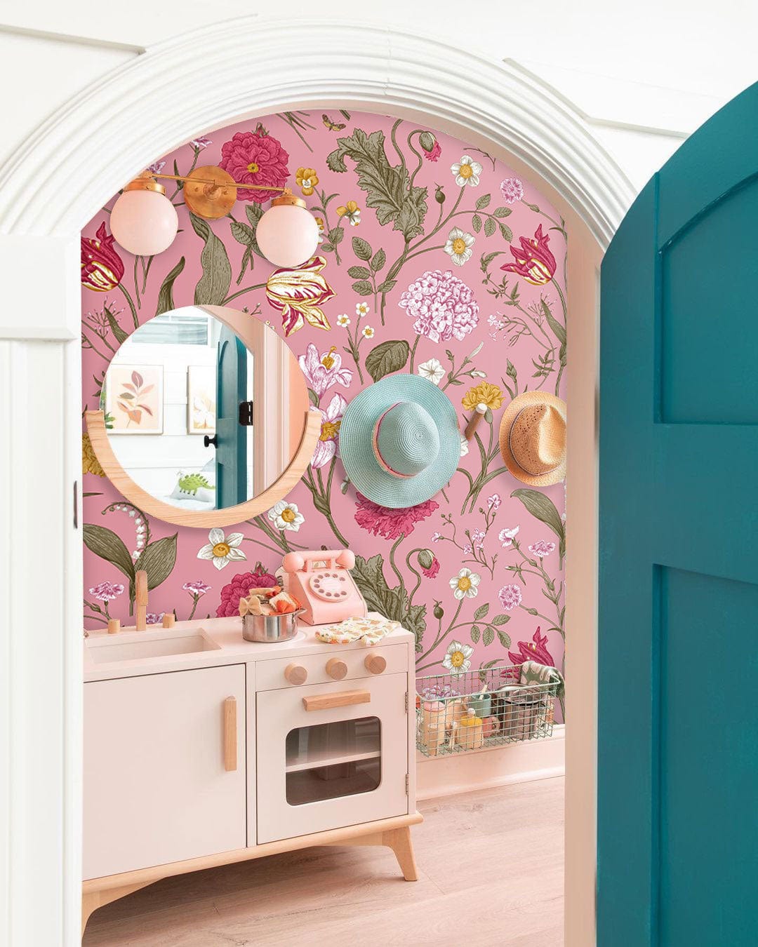 Playroom with pink floral wallpaper, natural wood play kitchen, colourful hats, arched doorway, round mirror.