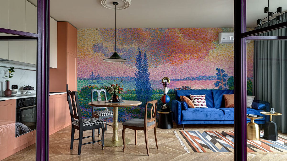 Vibrant pointillist landscape mural wallpaper in kitchen-living space with blue sofa, parquet flooring, eclectic chairs.