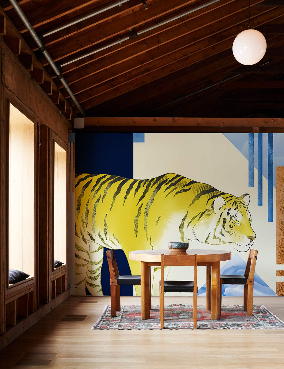 Golden Tiger geometric mural wallpaper, blue and yellow hues, wood table, chairs, wooden ceiling, rug.