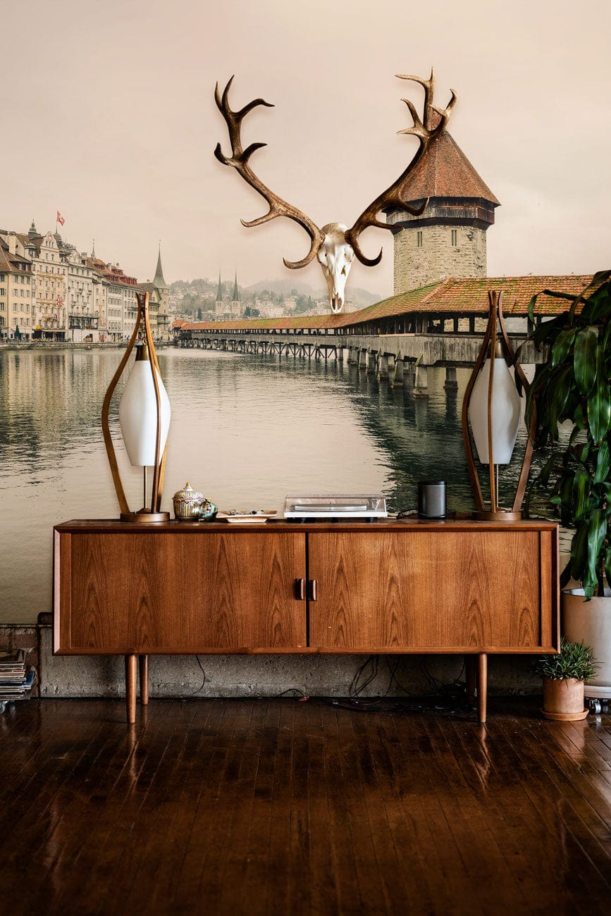 Vintage European lakeside mural wallpaper with wooden console, antlers decor, and dark wooden flooring.