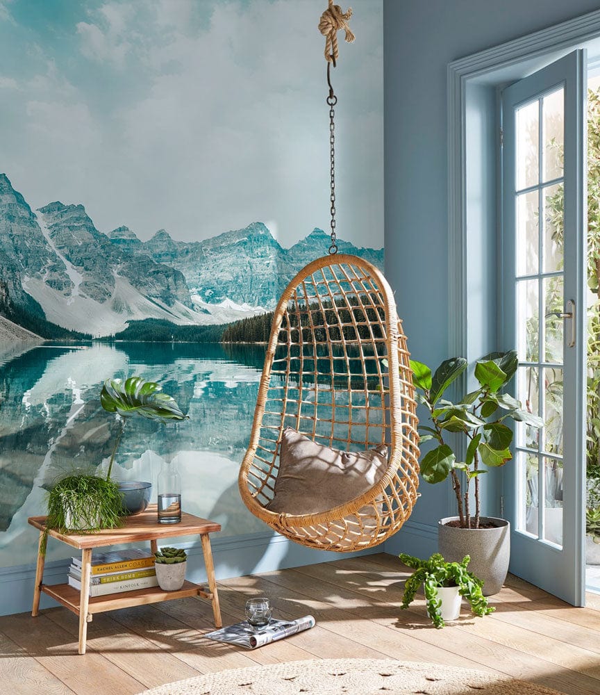 Serene Teal Mountain Lake Mural in a cosy lounge; wicker chair, light wood flooring, glass doors.