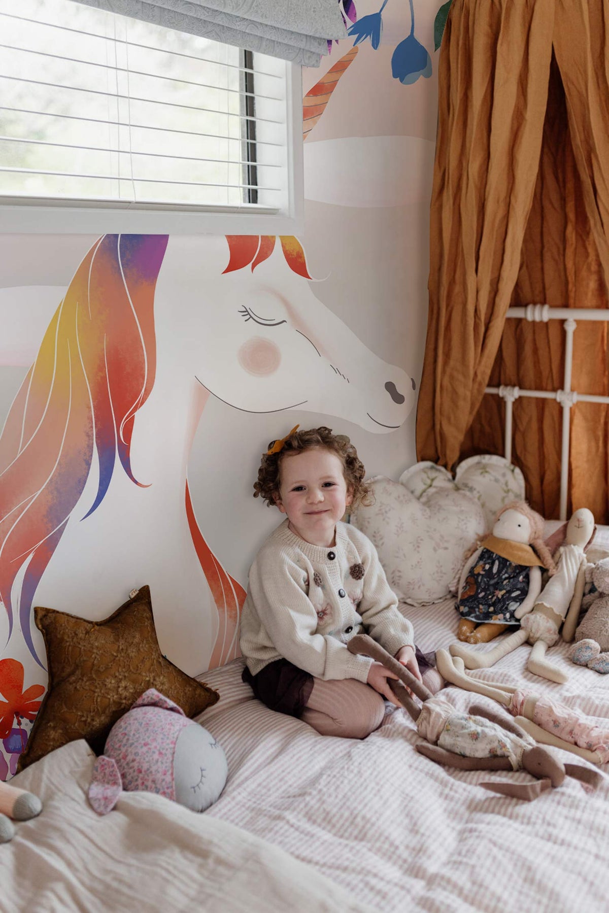 Child's bedroom with whimsical rainbow unicorn mural wallpaper, colourful bedding, and soft toys on bed.