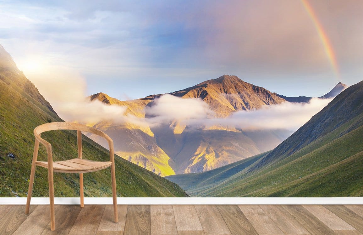 Serene mountain landscape mural wallpaper with rainbow and golden glow, featuring wooden chair on wooden flooring.