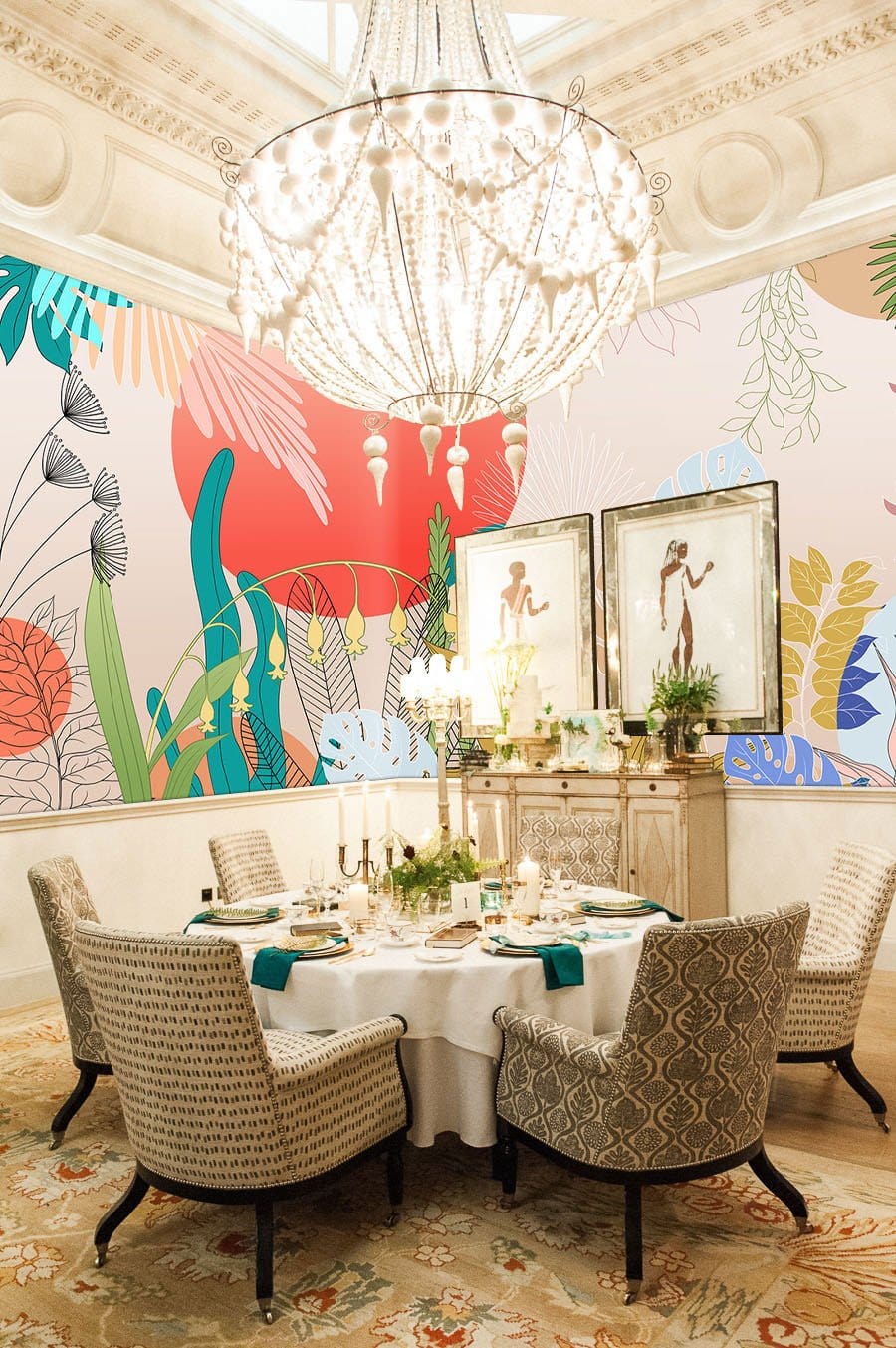 Tropical mural wallpaper in dining room, featuring elegant chandelier, patterned chairs, and antique sideboard.