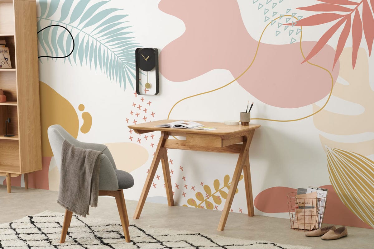 Tropical abstract botanical wallpaper mural, soft peach and sage tones, wooden desk, grey chair, rug.