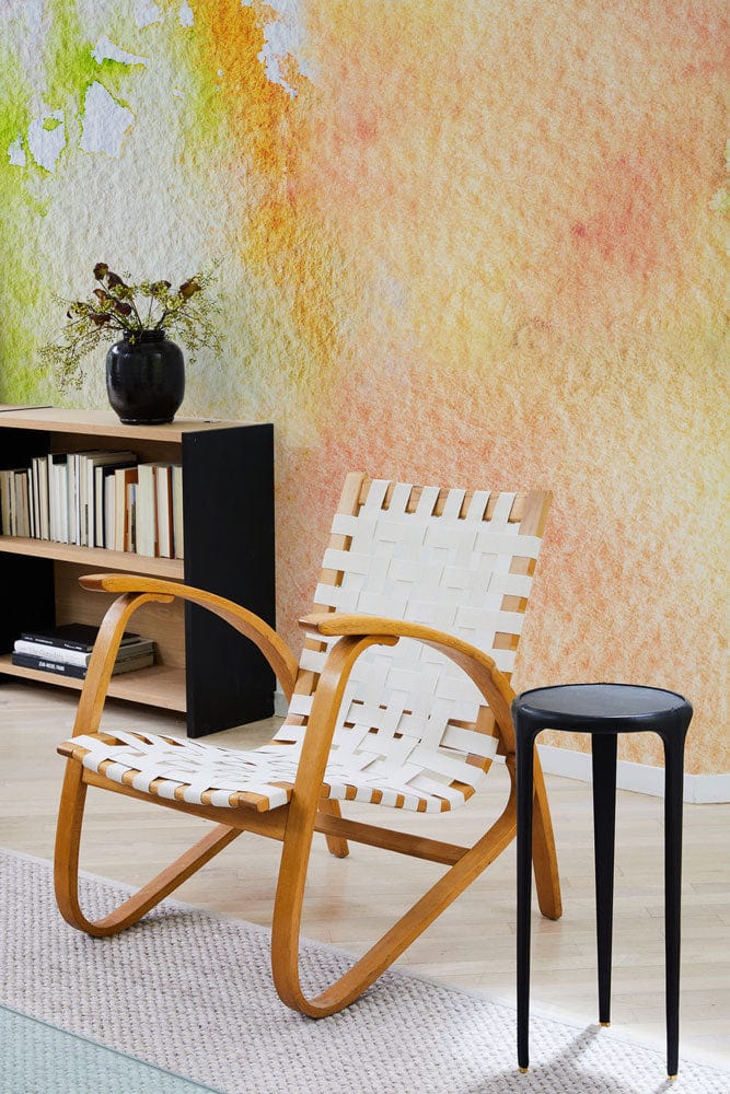 Soft pastel watercolour abstract mural wallpaper, woven chair, wooden shelves, black side table, light flooring.
