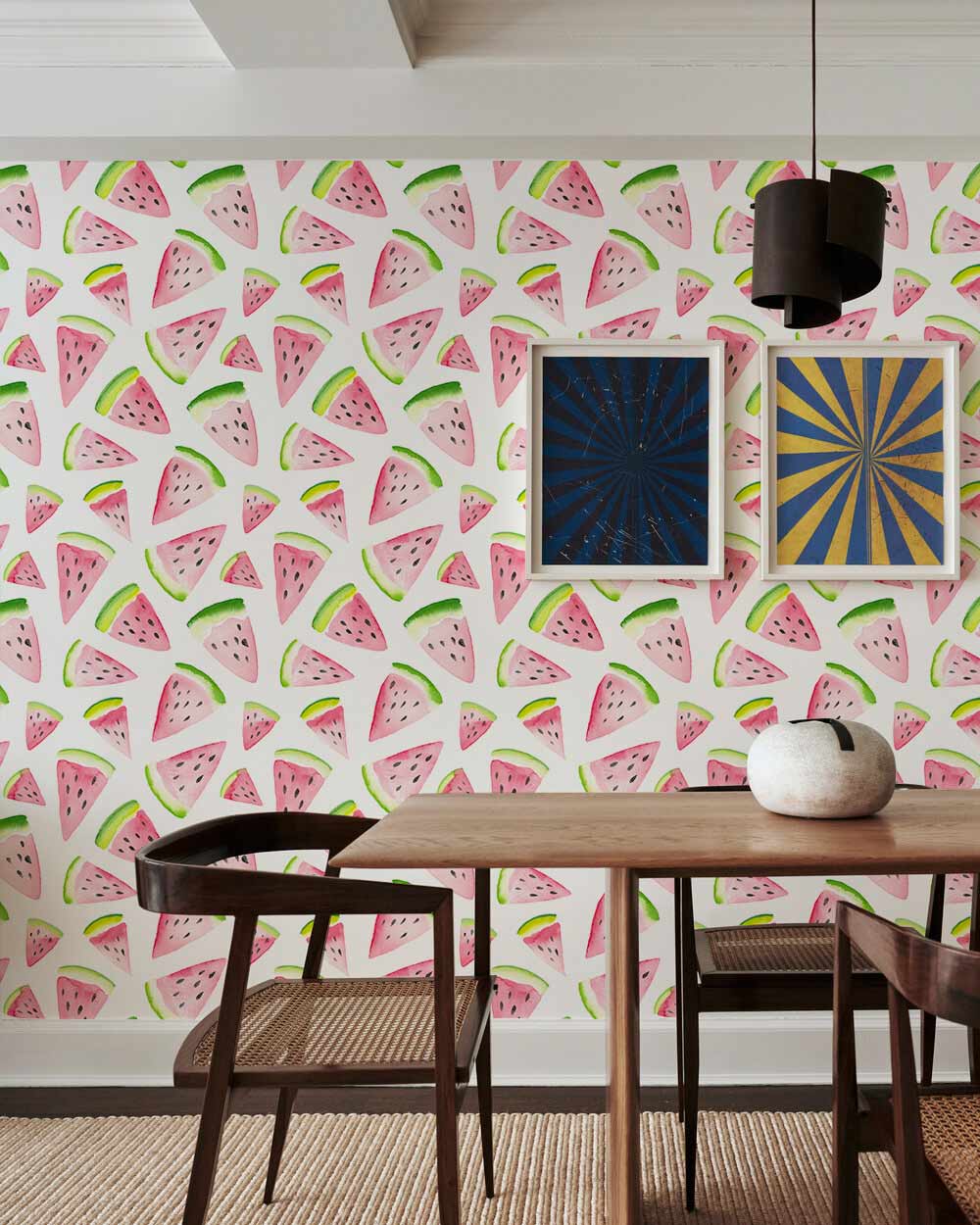 Dining room with vibrant watermelon-patterned wallpaper, wooden table, woven chairs, graphic art frames.