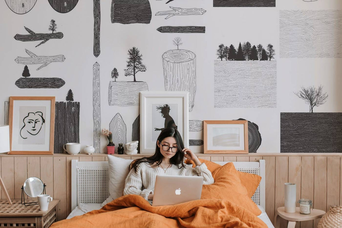Woman in cosy bedroom with Monochrome Woodland Sketch Mural Wallpaper, rustic wood panelling, and orange bedding.
